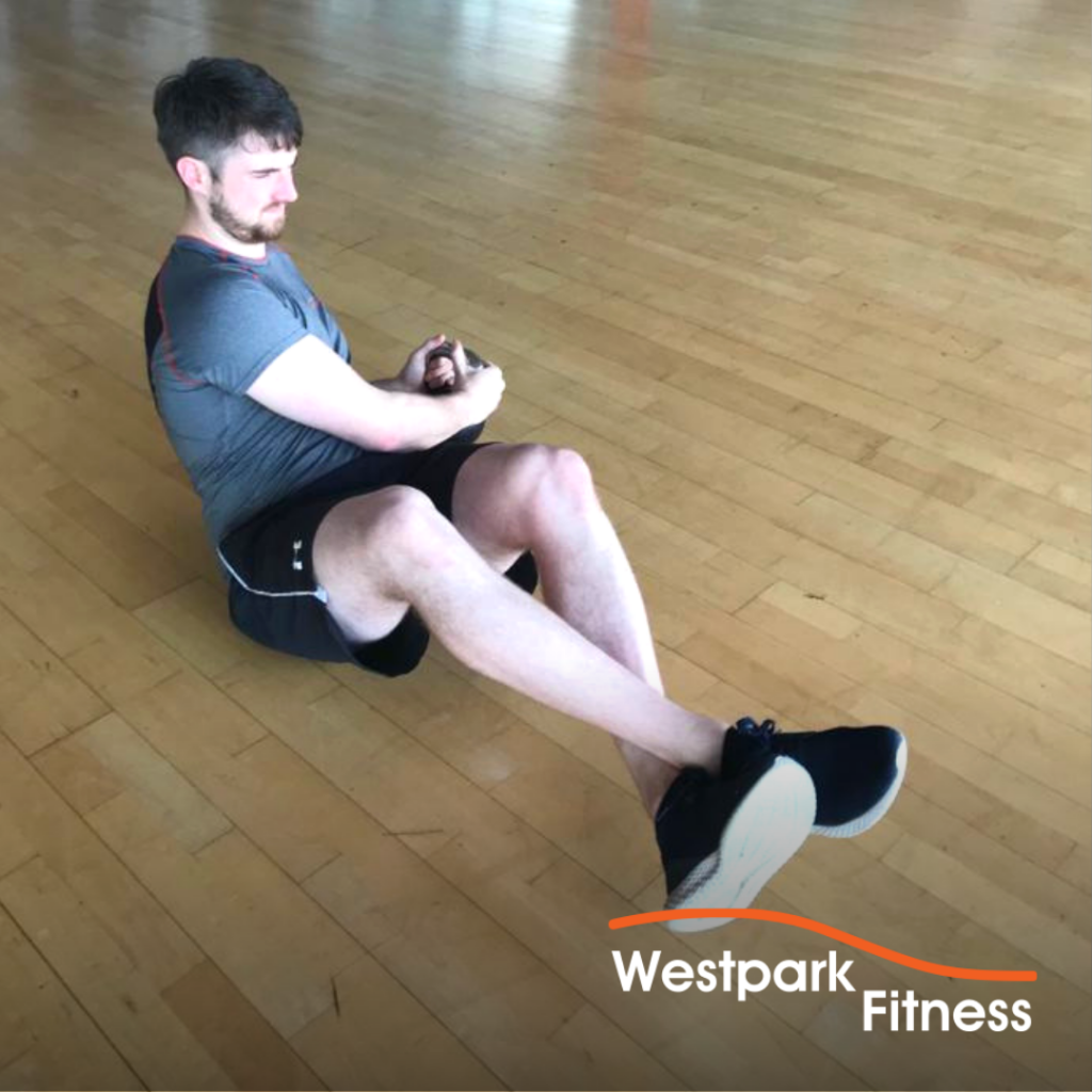 kettlebell russian twsit exercise being completed by a male gym goer lying on a wooden floor with a kettlebell gripped by both hands